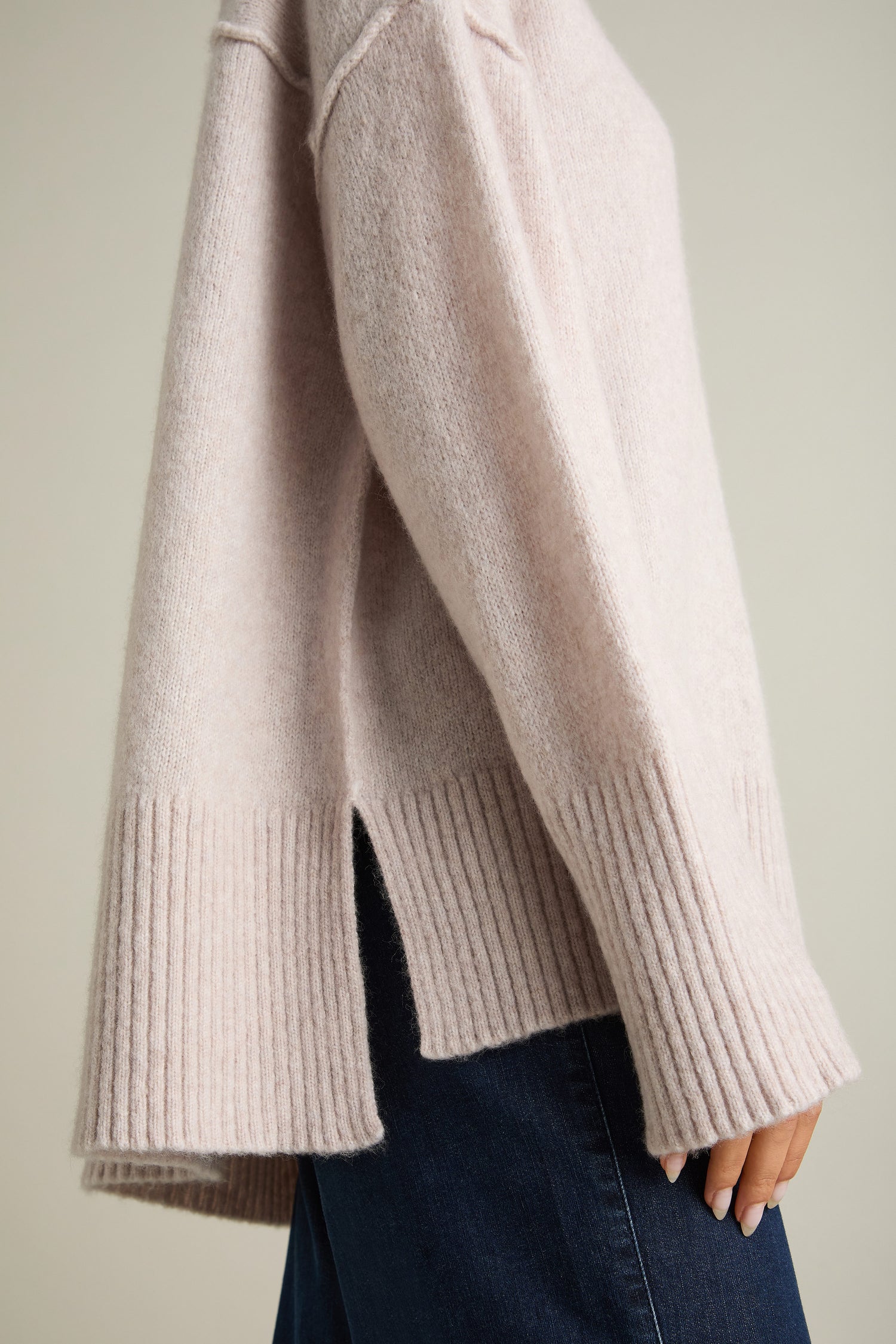 Close-up of a person wearing a Charming Merino Polo Jumper in light beige, showcasing ribbed cuffs and hem, intricate cable knit detailing, and a side slit. The person is also wearing blue denim jeans.