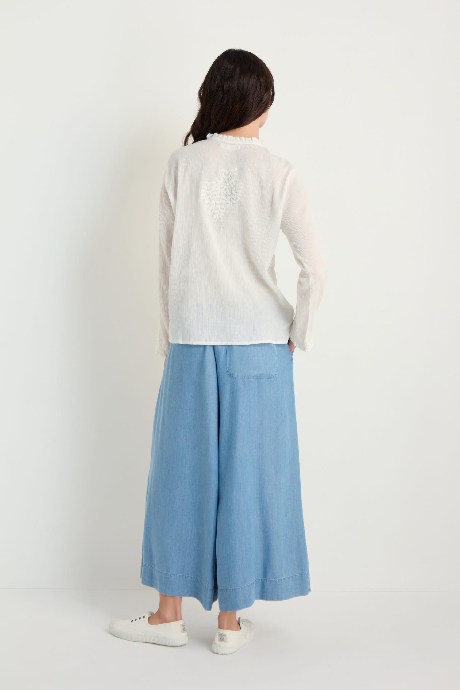 A person stands with their back to the camera, wearing the Embroidered Cotton Top in natural white, paired with light blue wide-leg pants and white sneakers, set against a plain white background.