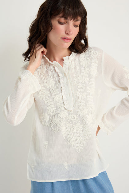 A woman wears the Embroidered Cotton Top in natural white, featuring sheer long sleeves, paired with light blue jeans, standing against a plain background.