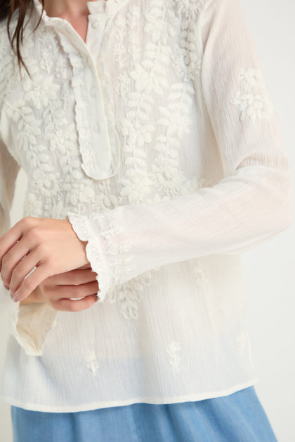 A person wearing the Embroidered Cotton Top in natural white—a sheer, long-sleeved women’s blouse with elegant embroidery and partial button placket—paired with a blue skirt, shown from shoulders to hips.