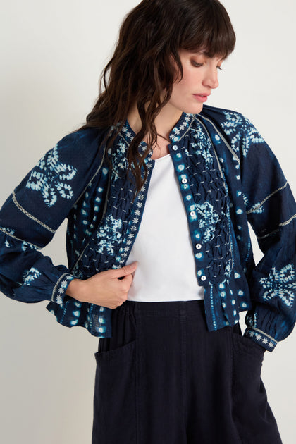 A woman wearing the Mixed Shibori Shirt, a dark navy patterned jacket with embroidered trim, and dark high-waisted pants stands with one hand in her pocket, looking to the side.