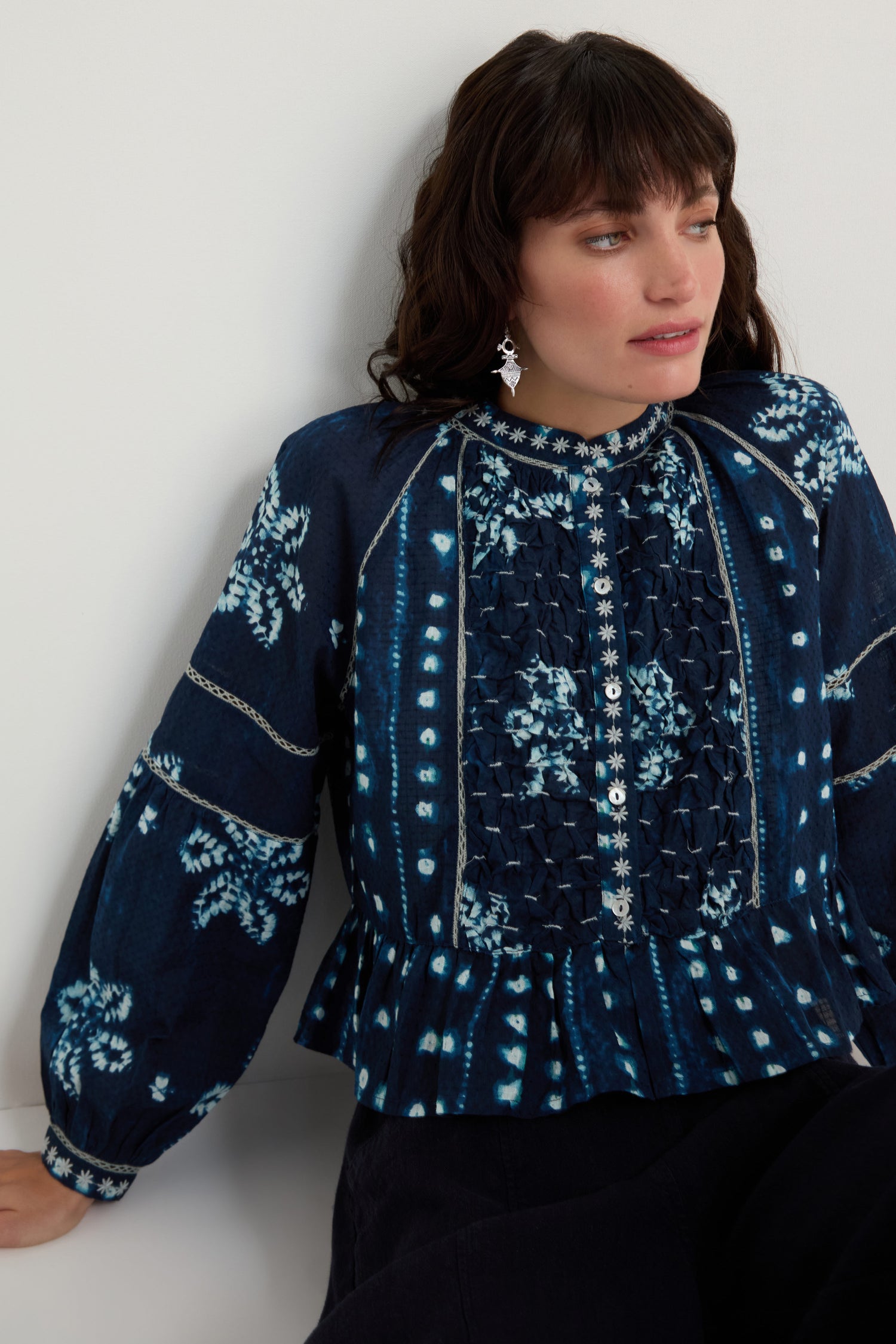 Woman wearing the Mixed Shibori Shirt—dark blue with white floral and dot patterns, pure cotton, long sleeves, embroidered trim—sitting against a white background.