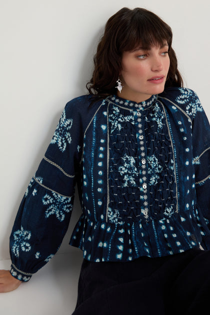 Woman wearing the Mixed Shibori Shirt—dark blue with white floral and dot patterns, pure cotton, long sleeves, embroidered trim—sitting against a white background.