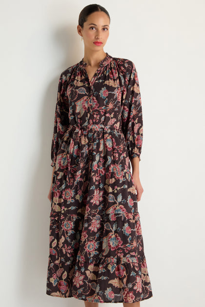A woman wears the Iris Print Tiered Dress, featuring long sleeves and an ankle-length black floral and paisley print, posing against a plain white background.