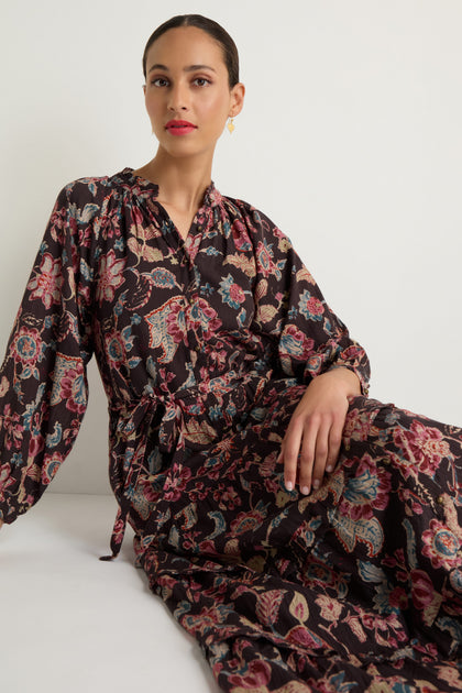 Woman sits against a plain white background wearing the Iris Print Tiered Dress, a long-sleeved dress featuring floral and paisley prints with a tie waist.