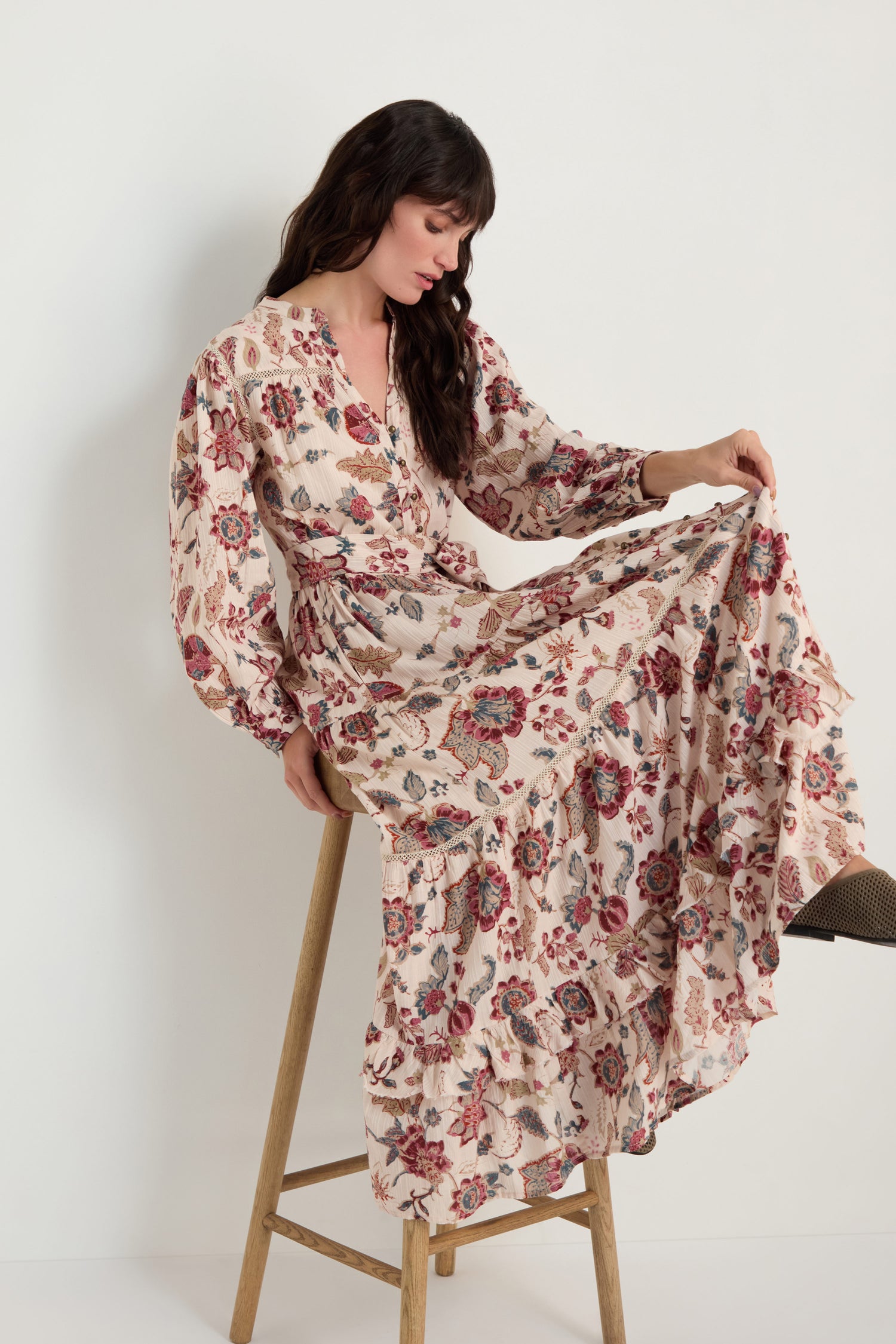 A woman sits on a wooden stool wearing the Floral Button-Through Tiered Dress, featuring pink and red flowers on a light background.