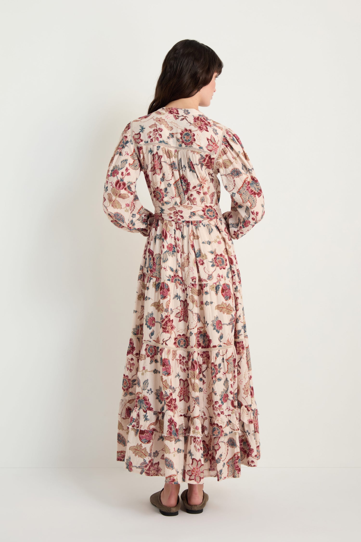 A woman faces away, wearing the Floral Button-Through Tiered Dress with long sleeves and flat shoes, set against a plain white background.