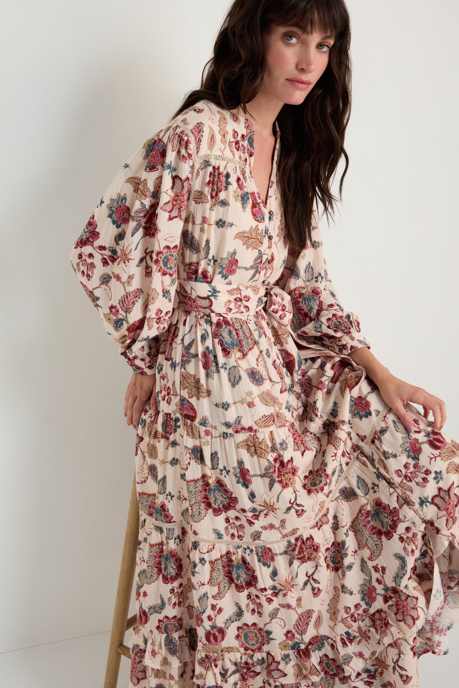 A woman sits against a plain light background, wearing the Floral Button-Through Tiered Dress—a long-sleeved, cream-colored dress with floral print, tiered design, belted waist, and button-through front.
