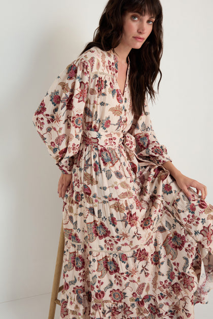 A woman sits against a plain light background, wearing the Floral Button-Through Tiered Dress—a long-sleeved, cream-colored dress with floral print, tiered design, belted waist, and button-through front.