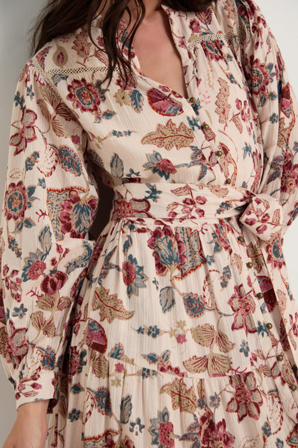 A woman wears the Floral Button-Through Tiered Dress featuring long sleeves, a belted waist, and floral layers in cream, red, and blue. Her face is partially out of frame.