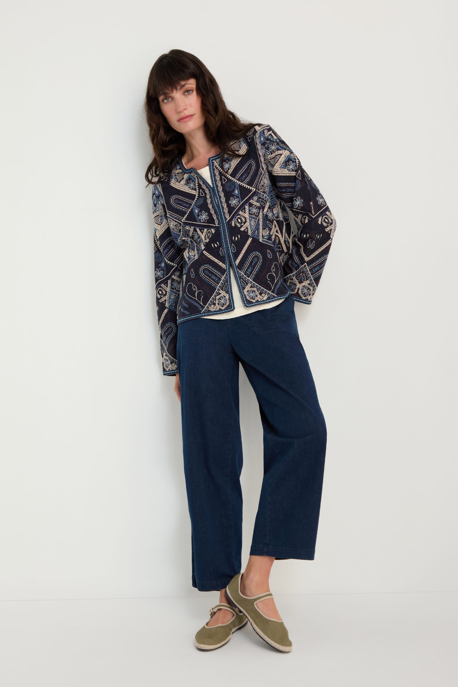 A woman stands against a white wall wearing the Embroidered Mosaic Jacket, styled with a white top, wide-leg blue jeans, and olive green Mary Jane shoes.