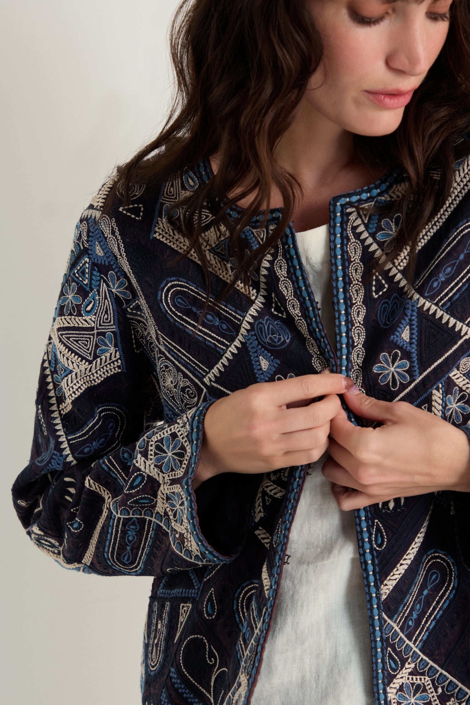 A woman adjusts the front of the Embroidered Mosaic Jacket, a boxy-fit piece with blue and beige geometric and floral embroidery, worn over a white top.