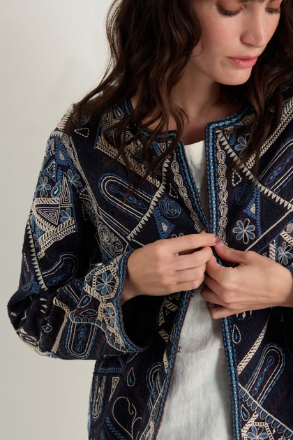 A woman adjusts the front of the Embroidered Mosaic Jacket, a boxy-fit piece with blue and beige geometric and floral embroidery, worn over a white top.