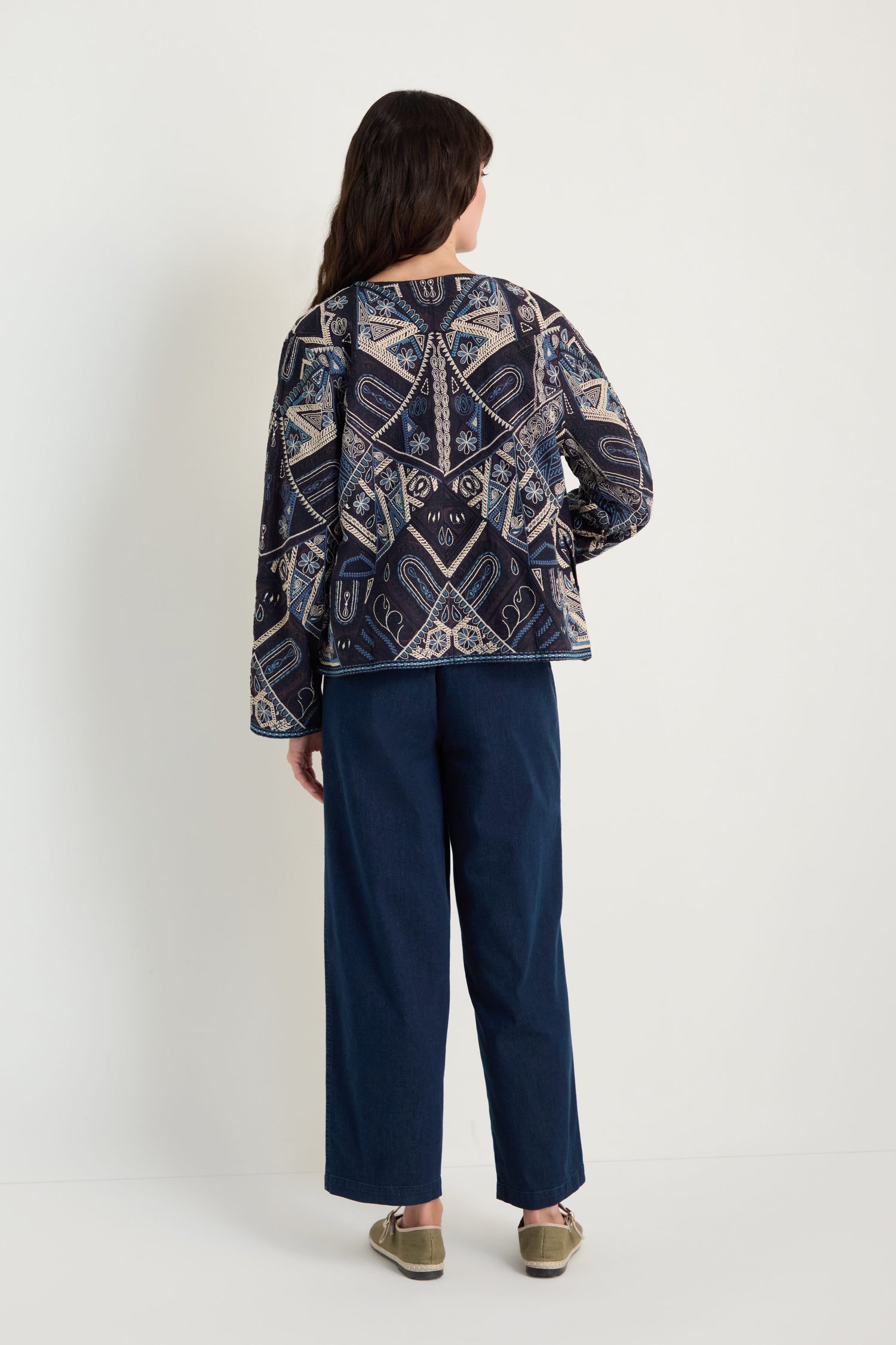 A person stands facing away, wearing the Embroidered Mosaic Jacket with a boxy fit and geometric and floral patterns, paired with wide-leg dark blue pants and gold slip-on shoes against a plain light background.
