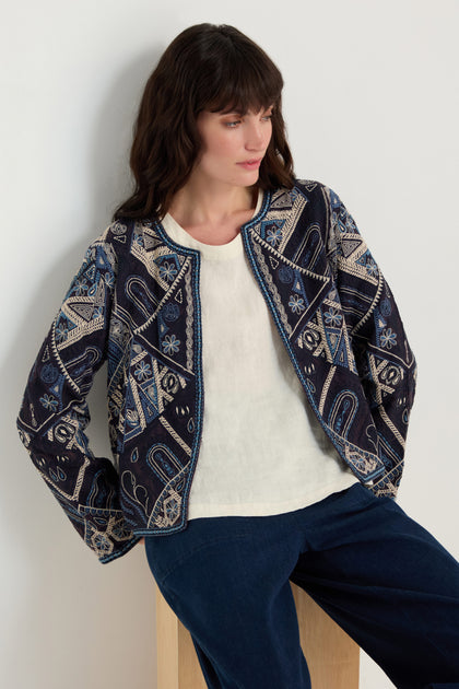 A woman with long dark hair wears the Embroidered Mosaic Jacket, featuring geometric and floral embroidery, paired with a white shirt and dark pants while sitting on a wooden stool against a plain light background.