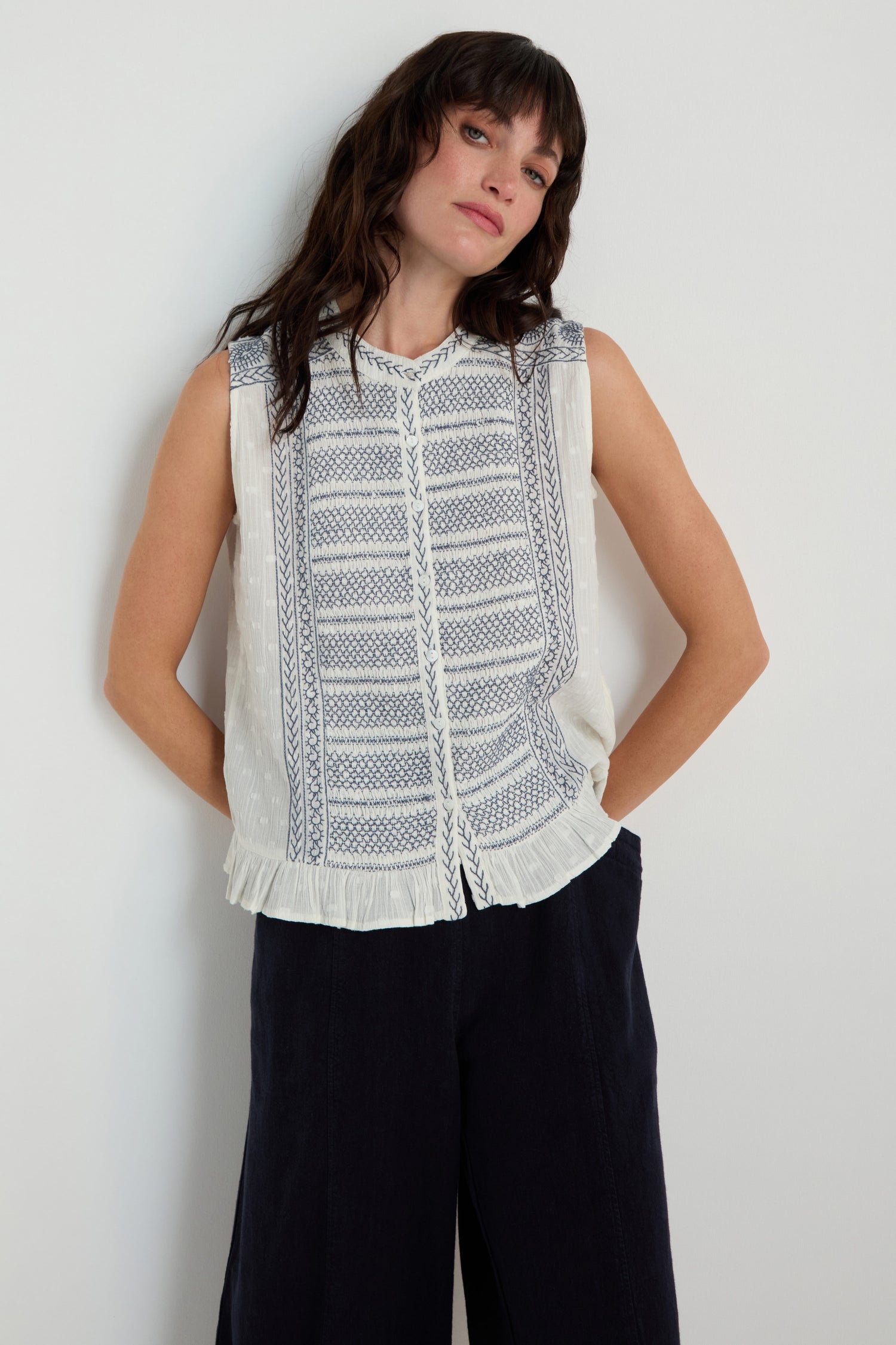 A woman stands against a plain wall wearing the Embroidered Button-Through Top with blue cross-stitch details and dark wide-leg pants.
