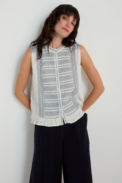 A woman stands against a plain wall wearing the Embroidered Button-Through Top with blue cross-stitch details and dark wide-leg pants.