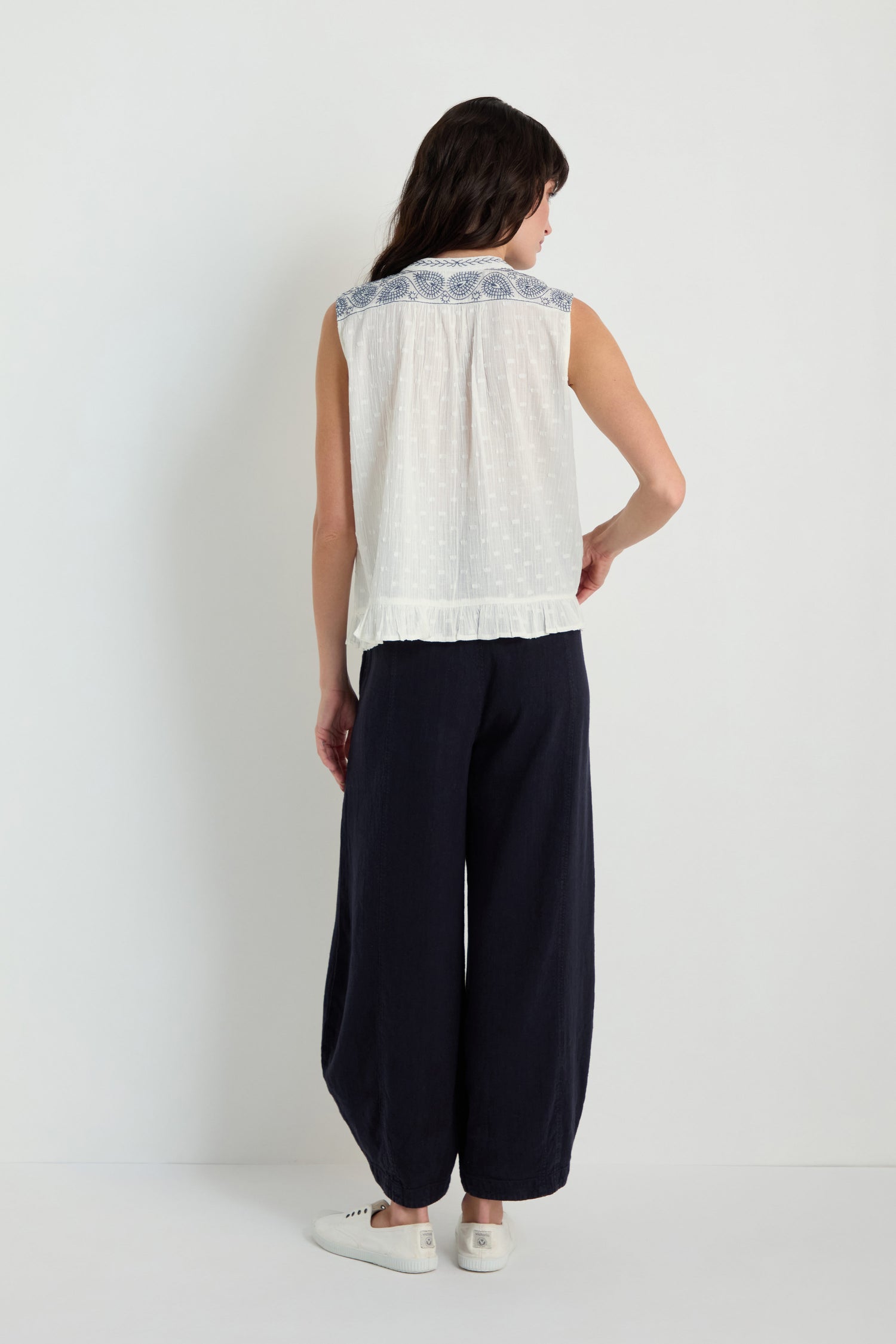A person stands facing away in an Embroidered Button-Through Top—white, sleeveless, with blue details—paired with wide-leg navy pants and white sneakers against a plain background.