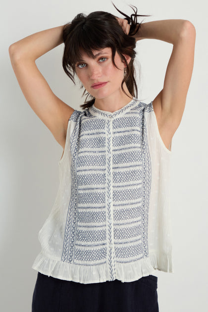 A woman stands with her hands behind her head, wearing the Embroidered Button-Through Top featuring blue patterns and a ruffled hem, set against a plain background.