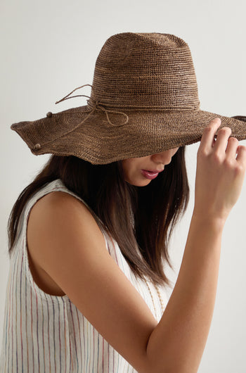 Woman wearing a sleeveless striped top and a Hand Woven Double Crochet Raffia Hat made from Madagascan raffia, holding the wide brim and facing slightly downward against a plain background.