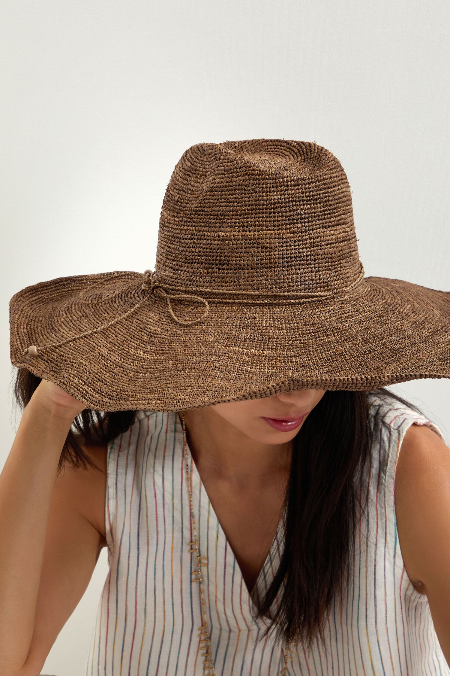 A woman wearing a Hand Woven Double Crochet Raffia Hat and a sleeveless, striped top sits against a plain white background.
