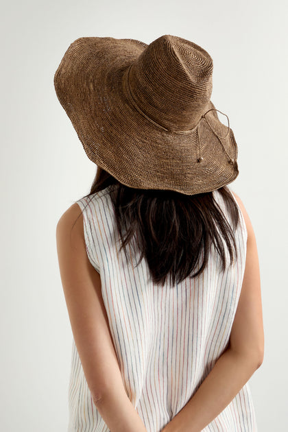 A person with long dark hair is seen from behind, wearing a Hand Woven Double Crochet Raffia Hat with an adjustable fit, and a sleeveless, light-colored striped top.