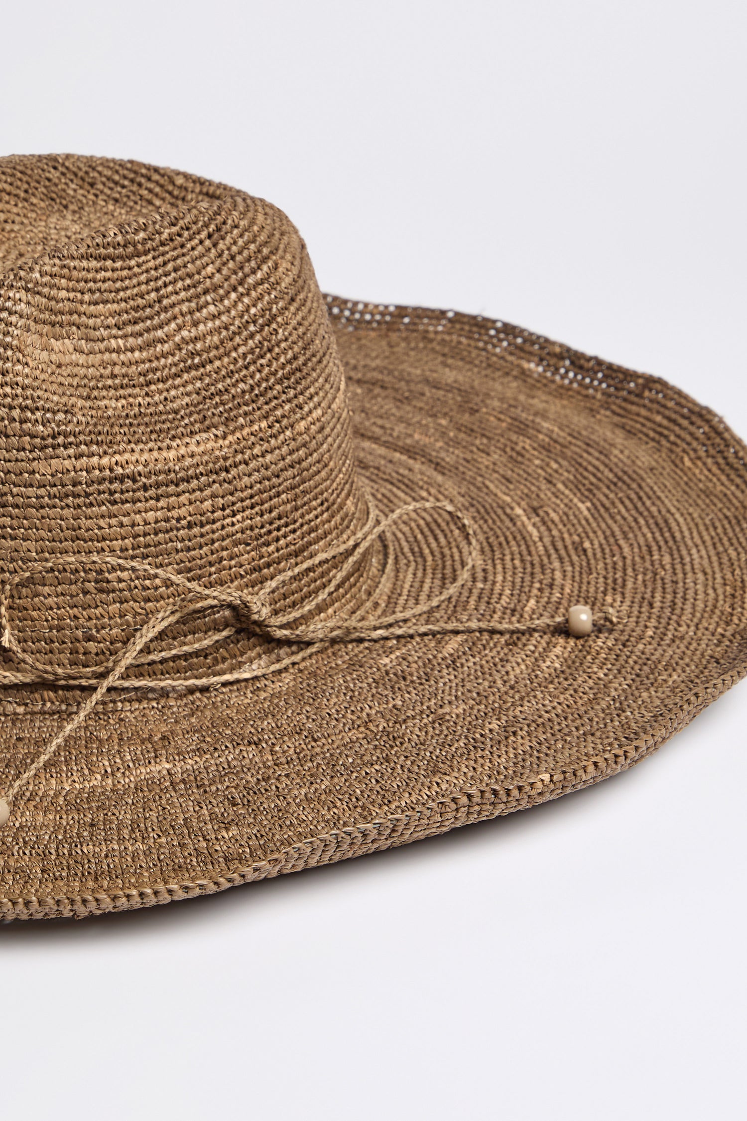 A close-up of the Hand Woven Double Crochet Raffia Hat made from Madagascan raffia, featuring a decorative string and bead detail and an adjustable fit, set against a plain background.