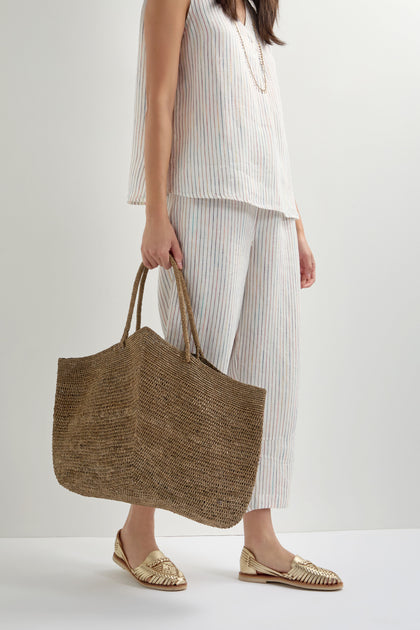 A woman holds the Large Handwoven Double Crochet Raffia Basket, pairing it with a sleeveless striped top, matching cropped pants, and gold woven flats—an effortless nod to slow fashion.