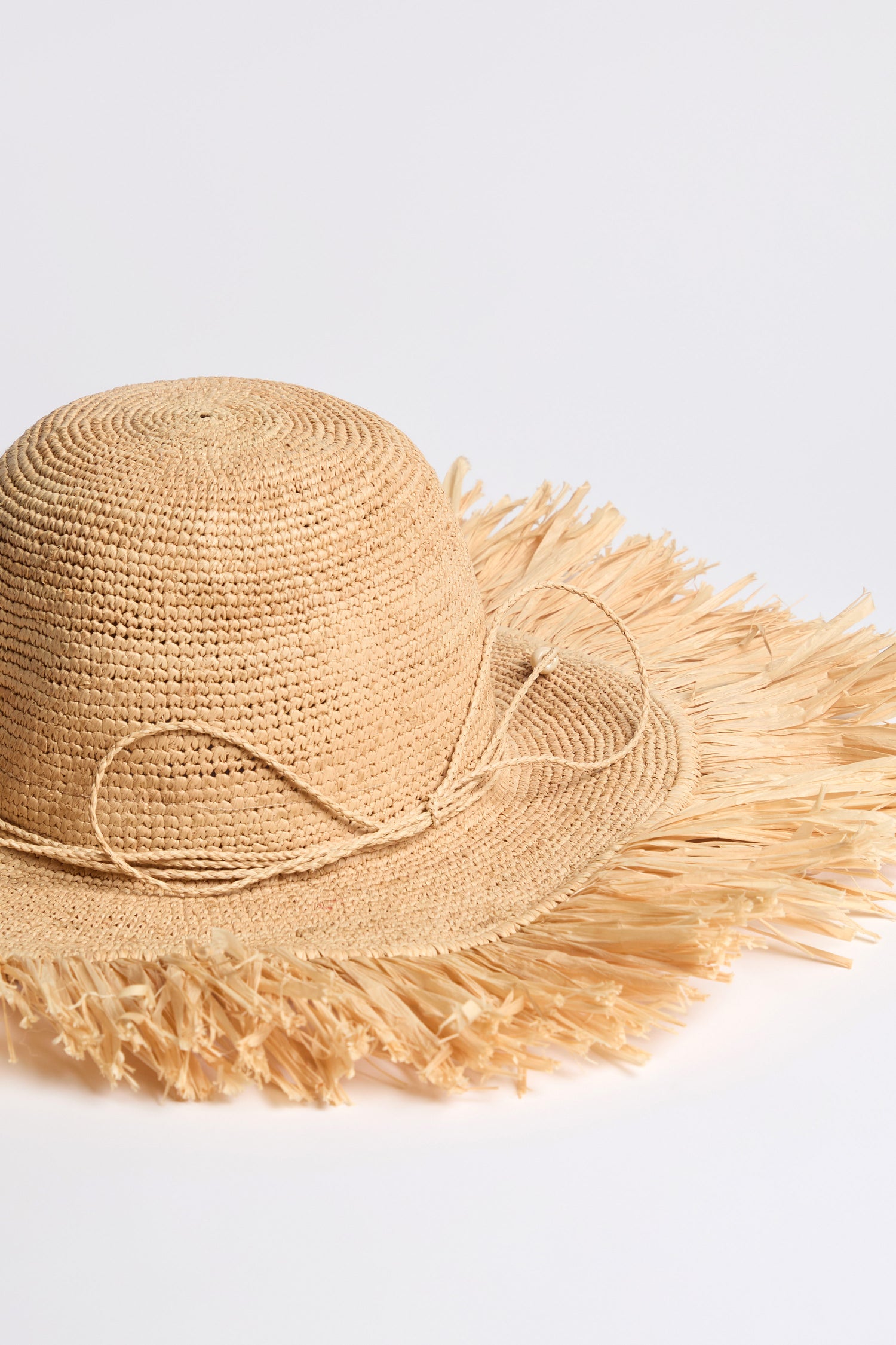 The Handmade Raffia Straw Fringe Hat features a round crown, frayed brim, and decorative bow. Crafted from sustainably harvested raffia, it's perfect for sunny holidays and shown against a plain white background.