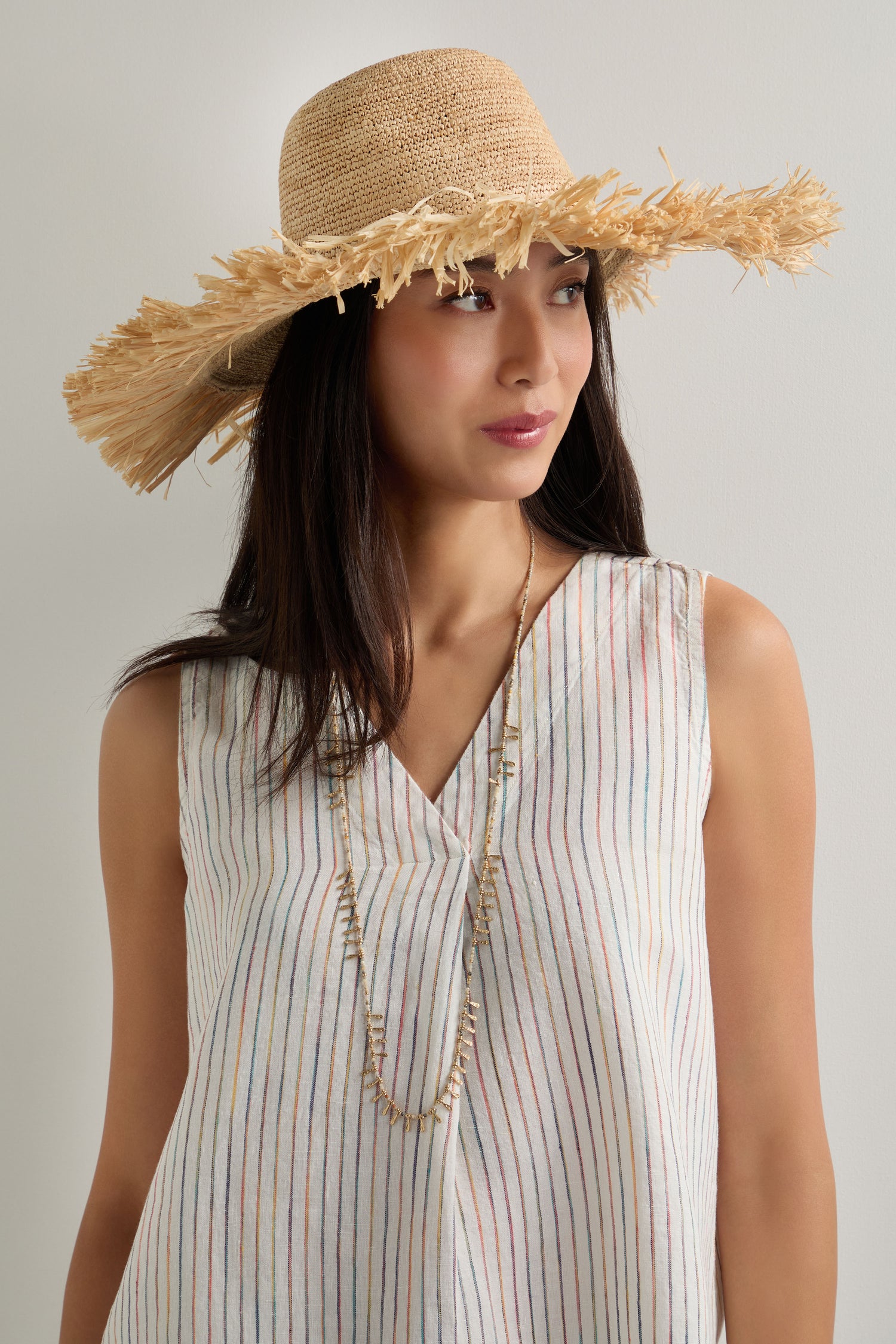Woman wearing a Handmade Raffia Straw Fringe Hat, a sleeveless striped top, and a long beaded necklace, standing against a plain light background.