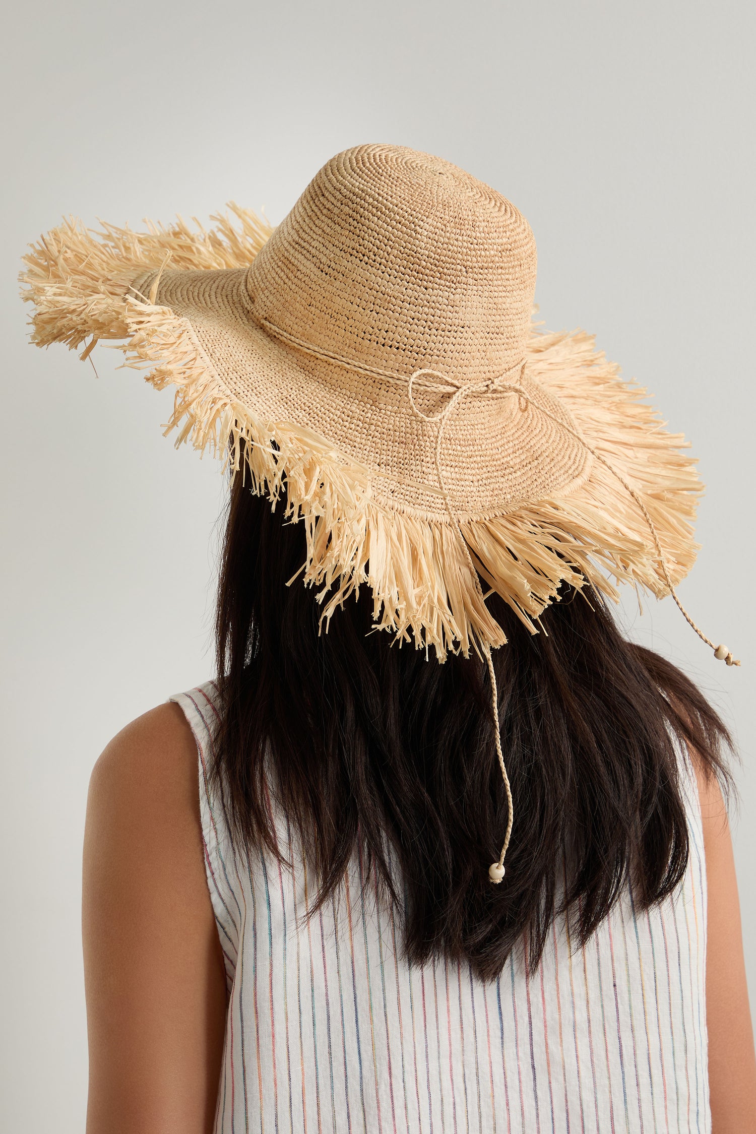 A person with long dark hair, seen from behind, wears a Handmade Raffia Straw Fringe Hat made from sustainably harvested raffia and a sleeveless, light-striped top.