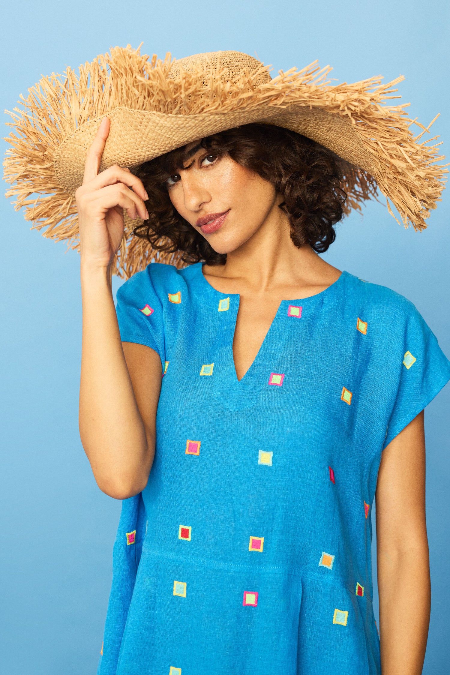 A woman in a blue dress with colorful squares poses against a blue background, wearing the Handmade Raffia Straw Fringe Hat and touching its brim with one hand.