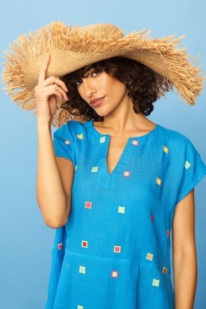 A woman in a blue dress with colorful squares poses against a blue background, wearing the Handmade Raffia Straw Fringe Hat and touching its brim with one hand.