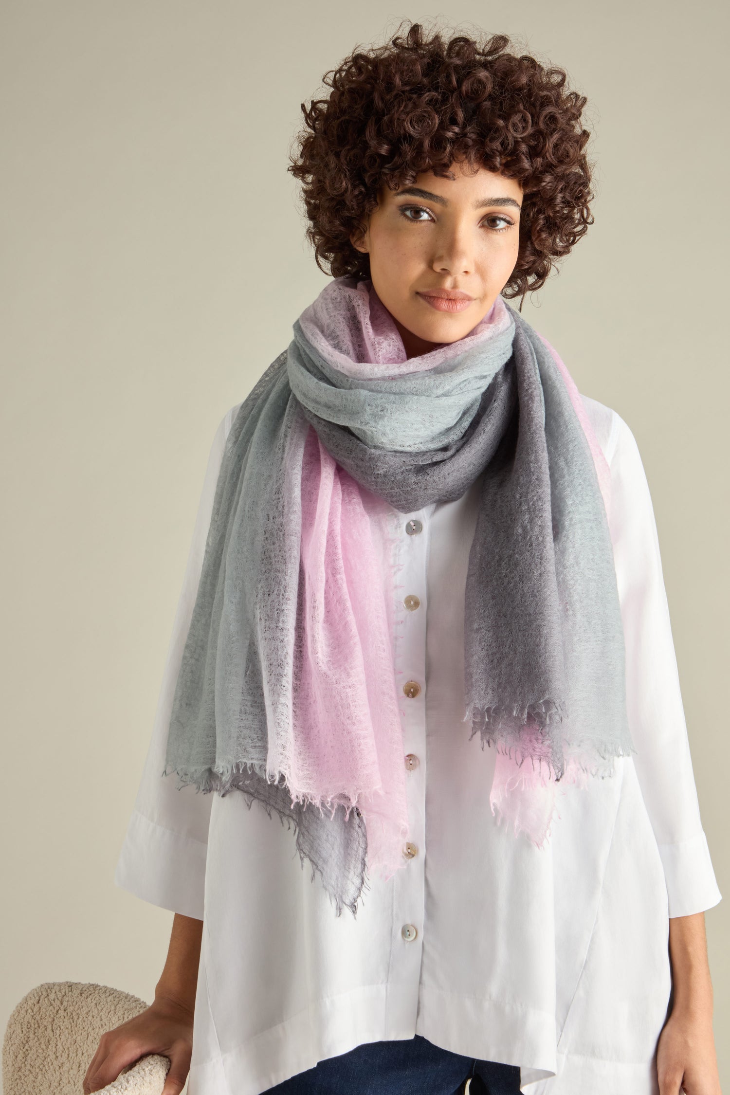 A person with curly hair, wearing a large gray and pink St Tropez Cashmere Shawl, handcrafted in Nepal, drapes it over a white button-up shirt while standing against a plain background.