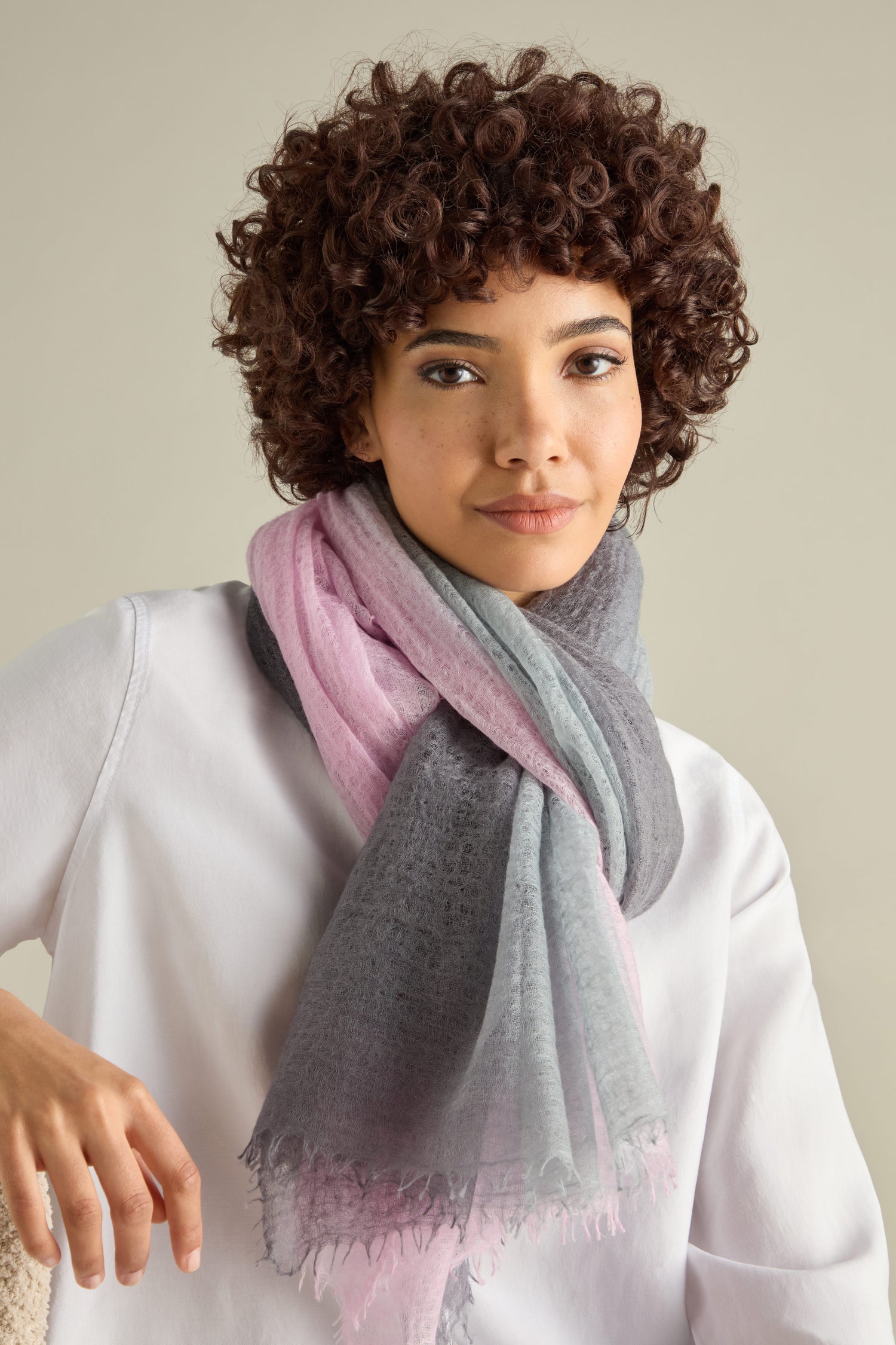 A person with curly hair in a white shirt drapes the St Tropez Cashmere Shawl, featuring pink and gray hues and handcrafted in Nepal, against a neutral background.