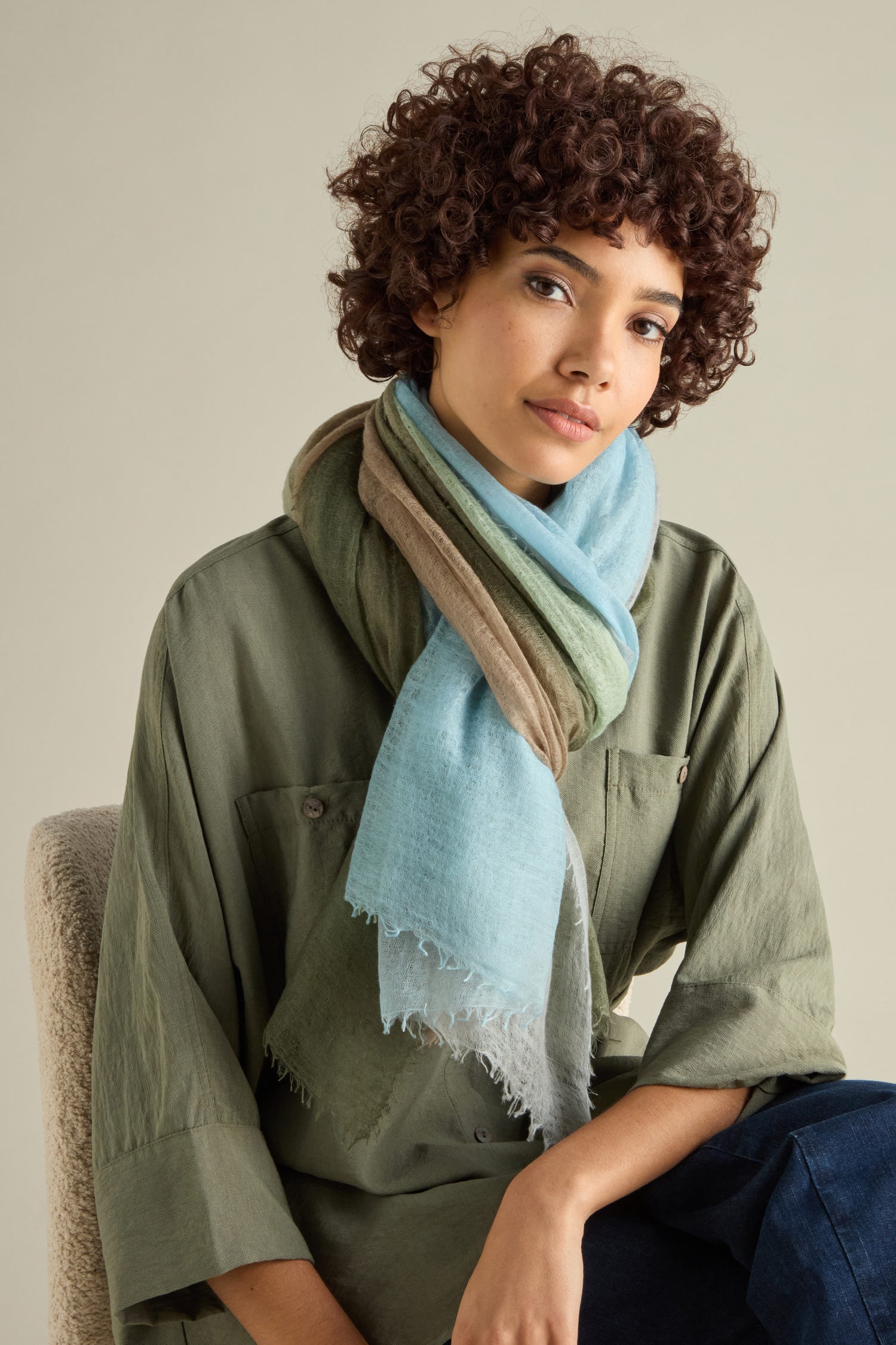 A person with curly hair in a green shirt and a layered St Tropez Cashmere Shawl sits on a chair against a neutral background, highlighting the beauty of items handcrafted in Nepal.