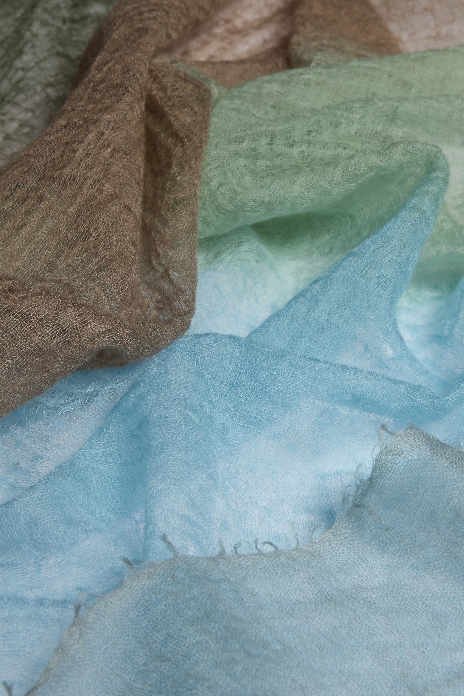 The St Tropez Cashmere Shawl features layered, sheer fabric transitioning from brown to green to blue earth tones. Handcrafted in Nepal, it embodies ethical practices and timeless elegance.