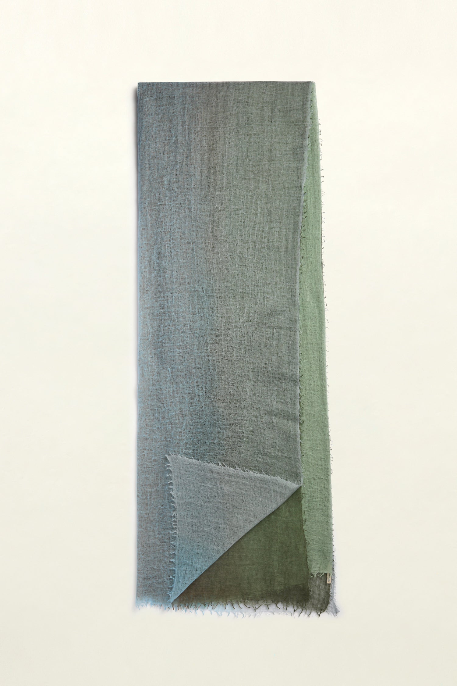 The St Tropez Cashmere Shawl is a long, rectangular, textured piece in shades of green and blue with frayed edges. It lies flat on a plain, light background and is handcrafted in Nepal using ethical practices, exuding both elegance and conscience.