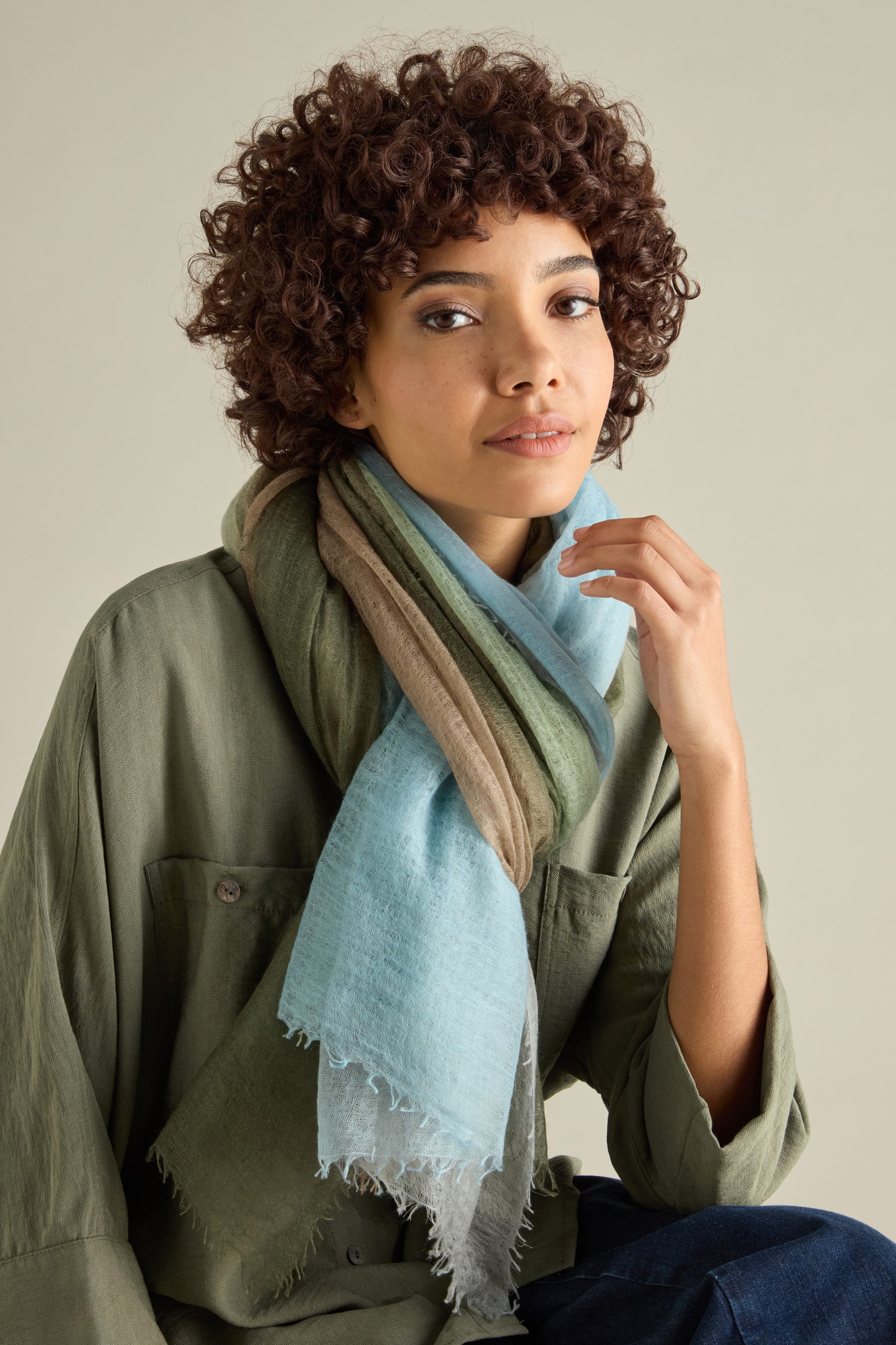 A person with curly hair wears a St Tropez Cashmere Shawl in green, beige, and light blue. Handcrafted in Nepal using ethical practices, they sit against a neutral background.