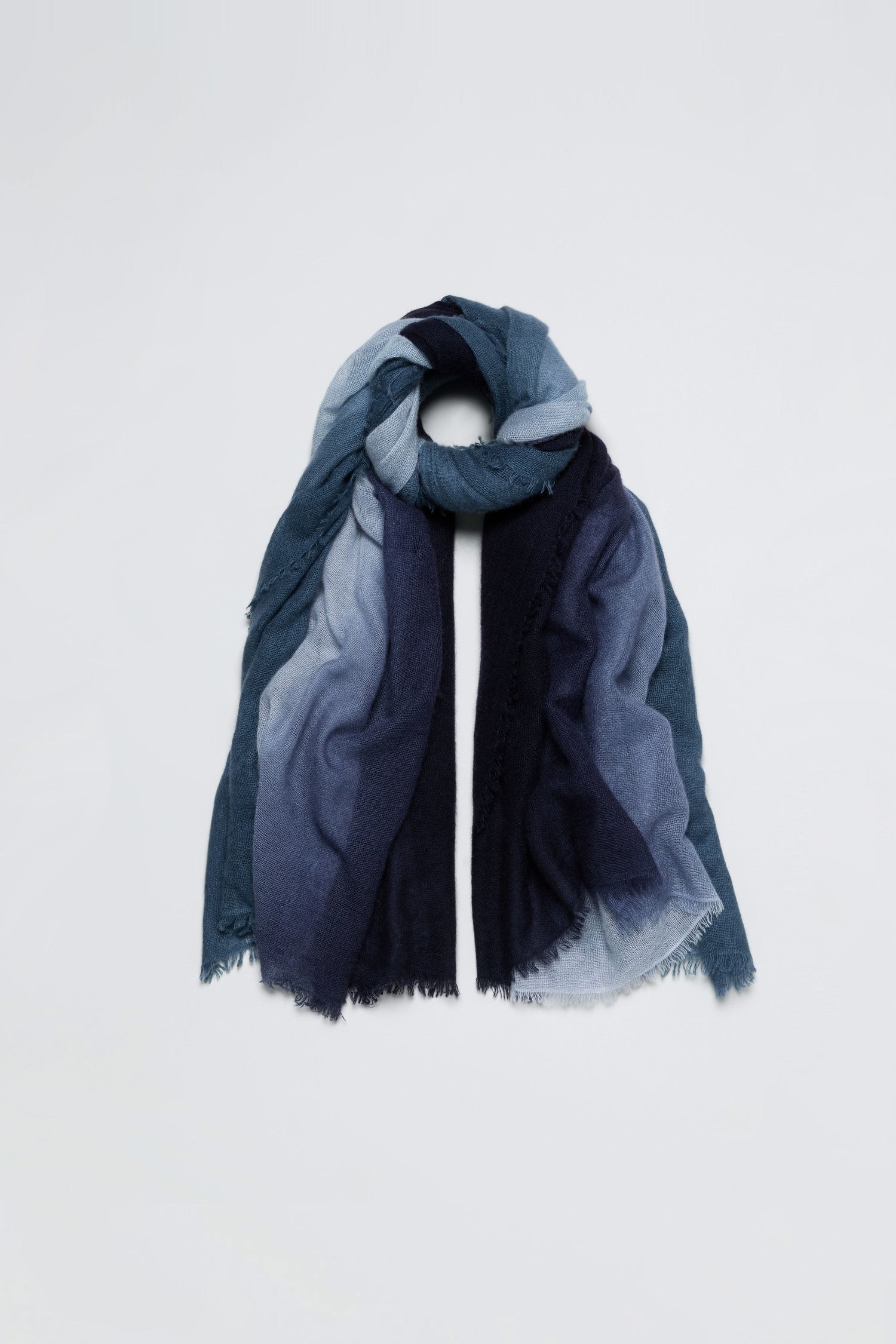 The Blue and Grey Soft Colourblend Cashmere Shawl, woven in Nepal, features a long rectangular shape with gradient blue tones and frayed edges, and is displayed in a loose loop on a plain white background.