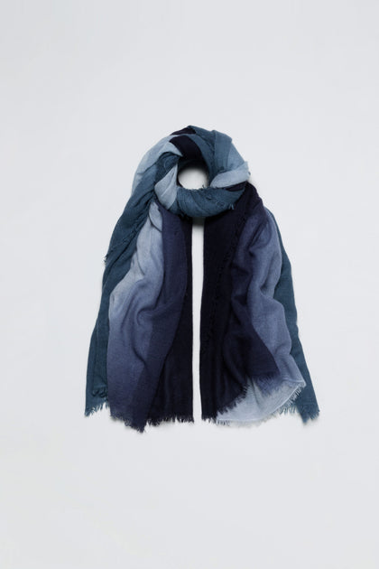 The Blue and Grey Soft Colourblend Cashmere Shawl, woven in Nepal, features a long rectangular shape with gradient blue tones and frayed edges, and is displayed in a loose loop on a plain white background.