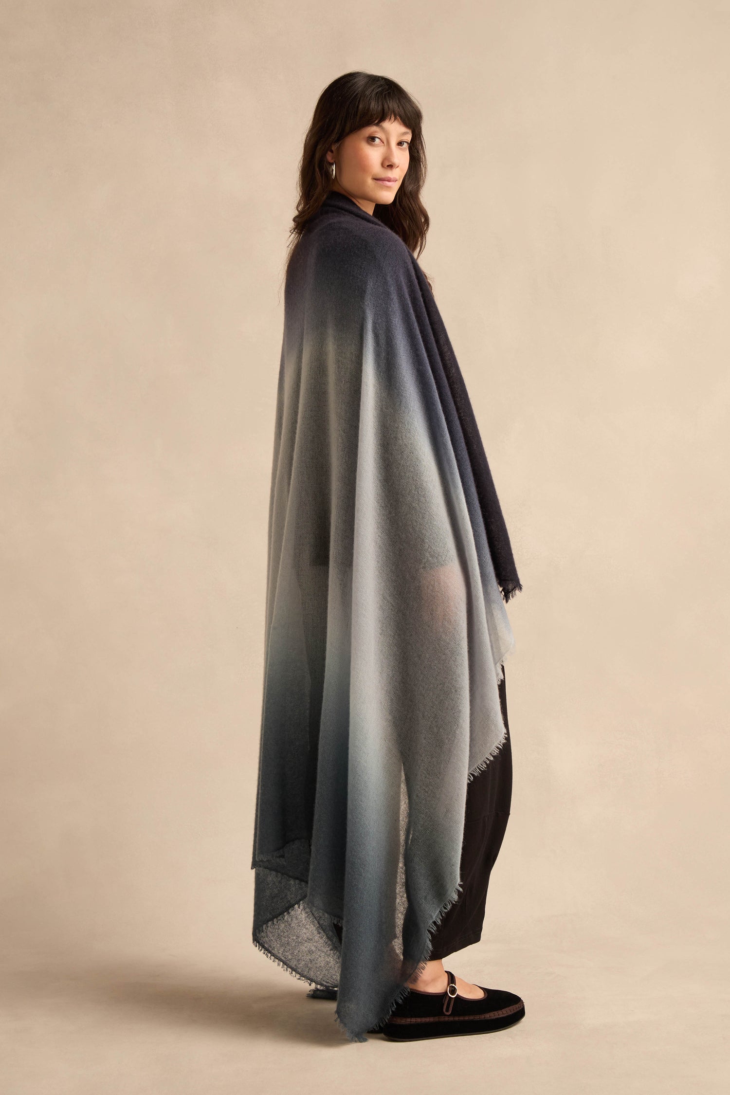 A person stands sideways against a plain beige background, wearing the Blue and Grey Soft Colourblend Cashmere Shawl—an ombré cashmere piece woven in Nepal in shades of dark blue to light gray—draped over black pants and black shoes.