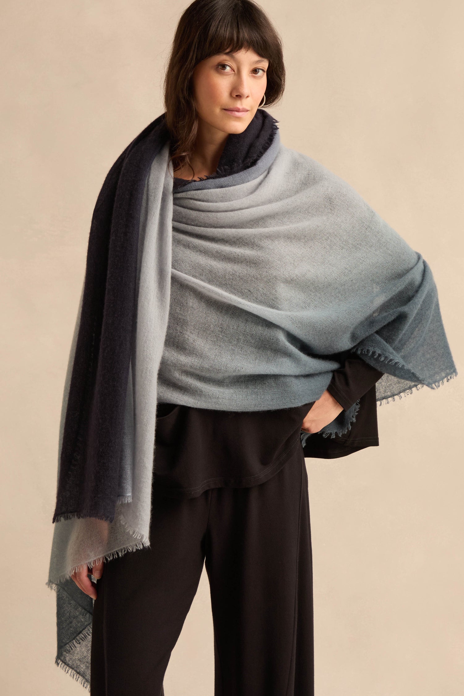 A woman stands against a neutral background, draping the Blue and Grey Soft Colourblend Cashmere Shawl over her shoulders. Paired with a black top and black pants, this luxurious shawl—expertly woven in Nepal—adds an elegant touch to her minimalist ensemble.