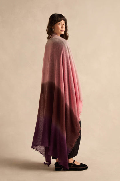 A woman in profile wears the Cherry and Pink Soft Colourblend Cashmere Shawl—woven in Nepal with pink, burgundy, and purple hues—draped over black pants and shoes, standing against a neutral background.