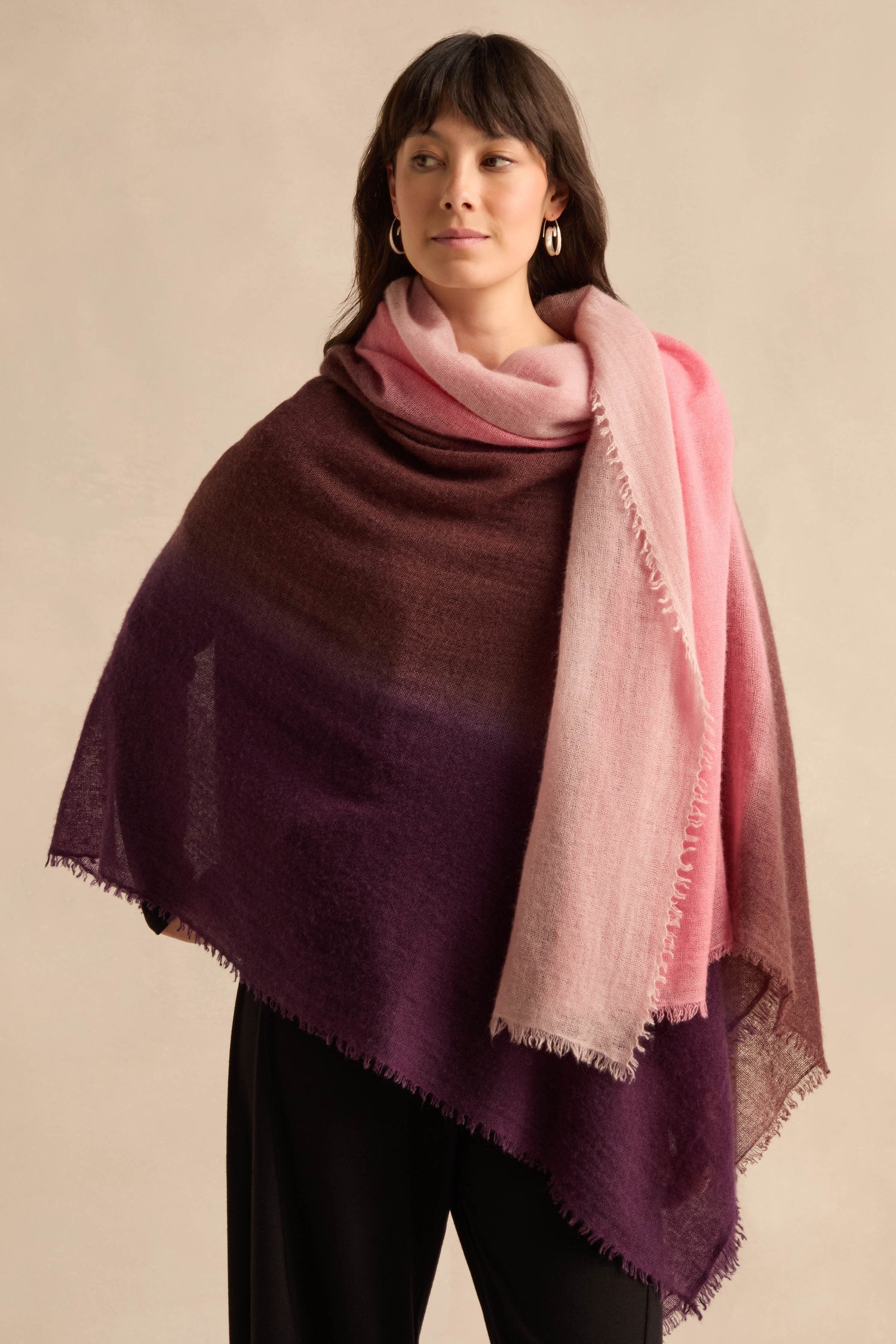 A woman stands against a neutral background, draped in the Cherry and Pink Soft Colourblend Cashmere Shawl, which features luxurious ombré hues of pink, brown, and purple over her black outfit. Expertly woven in Nepal, this elegant cashmere accessory adds a touch of sophistication.