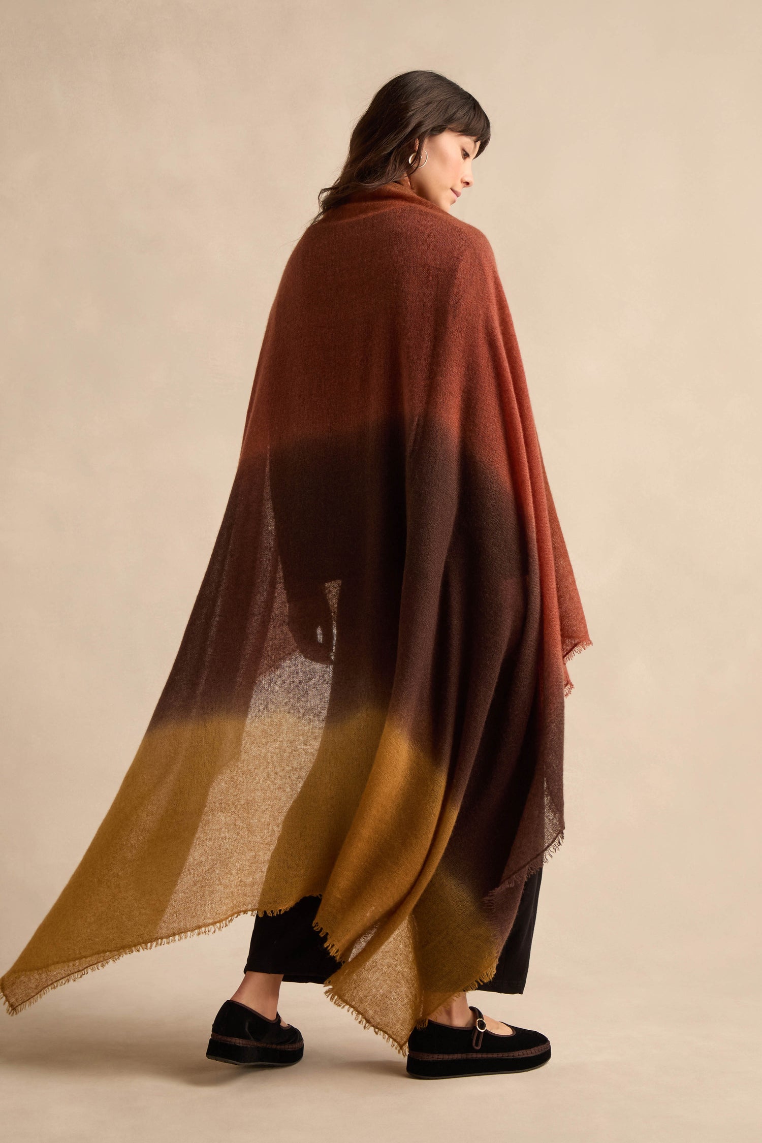 A woman is draped in the Rust and Ochre Soft Colourblend Cashmere Shawl—crafted in Nepal—which transitions beautifully from rust to dark brown to mustard yellow and features a delicate fringe trim. She pairs it with black trousers and flat shoes.
