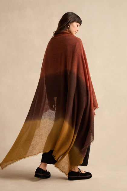 A woman is draped in the Rust and Ochre Soft Colourblend Cashmere Shawl—crafted in Nepal—which transitions beautifully from rust to dark brown to mustard yellow and features a delicate fringe trim. She pairs it with black trousers and flat shoes.