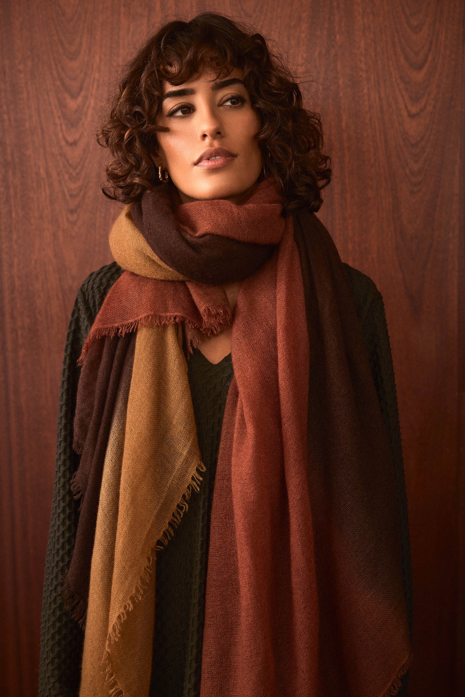 A person with short curly hair wears a textured dark sweater and the Rust and Ochre Soft Colourblend Cashmere Shawl—expertly woven in Nepal in rich brown, rust, and ochre tones—standing against a wooden background.