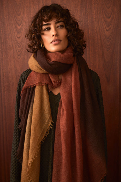 A person with short curly hair wears a textured dark sweater and the Rust and Ochre Soft Colourblend Cashmere Shawl—expertly woven in Nepal in rich brown, rust, and ochre tones—standing against a wooden background.