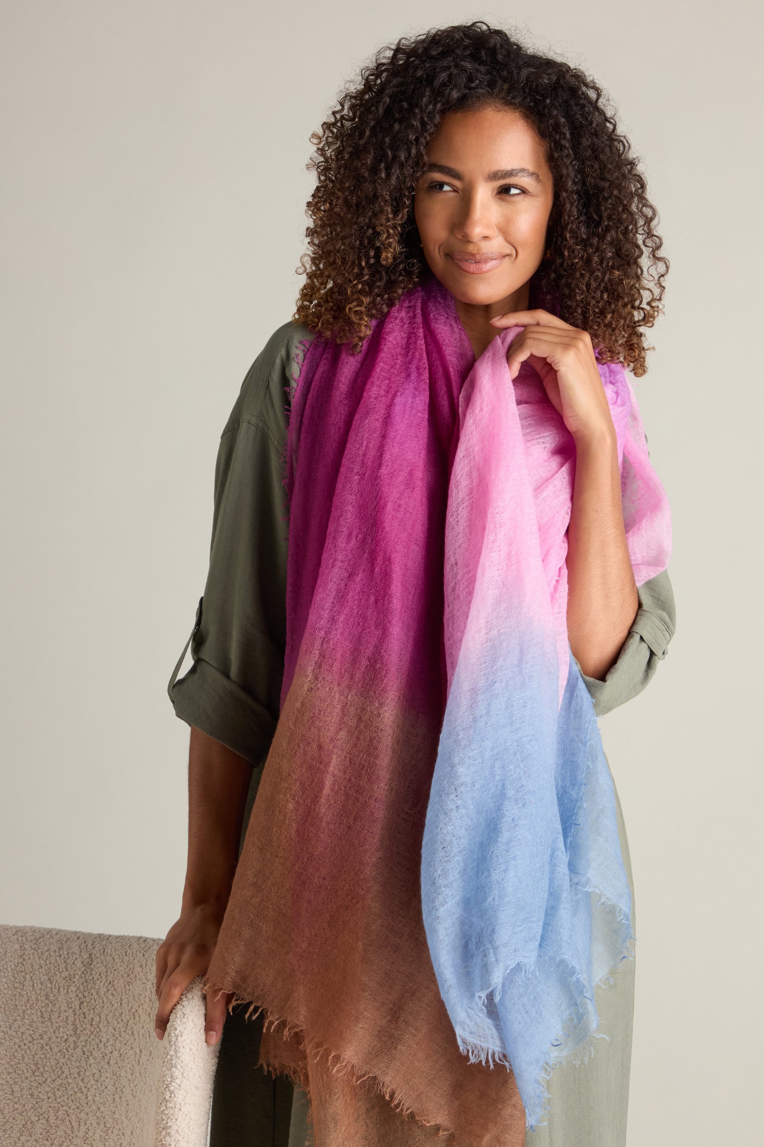 A person with curly hair wears a green dress and holds a luxurious accessory: the St Tropez Cashmere Shawl, showcasing a gradient watercolor design with shades of pink, brown, and blue.